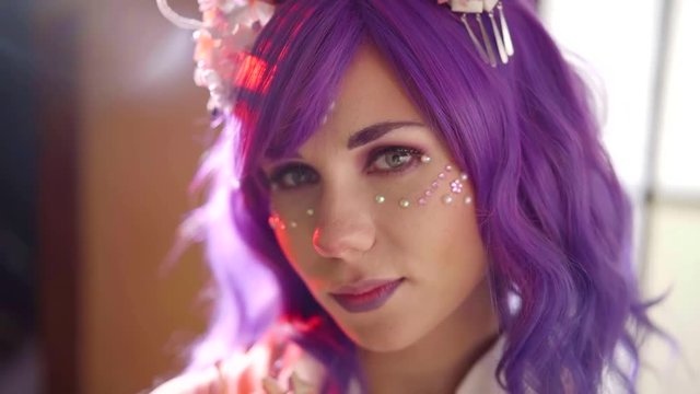 trendy violet haired girl with creative makeup with crystals on face is posing calmly in soft light