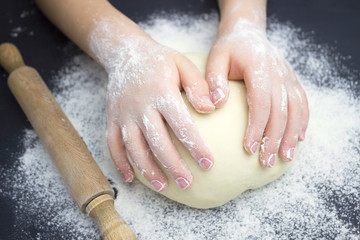 Kid's hands, some flour, wheat dough and rolling pin on the black table. Children hands making the rye dough for backing bread. Small hands kneading dough. Little child preparing dough for backing .