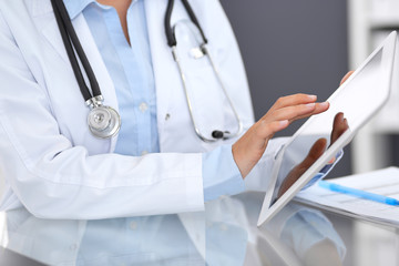 Close-up of female physician hands using digital tablet  while sitting at glass desk at hospital office. Medicine and healthcare concept