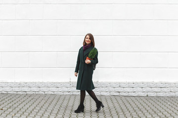 Beautiful young stylish woman wearing green coat, black high heel shoes and backpack walking through the city streets with small Christmas tree in her hands. New year concept. Christmas tree in a pot.