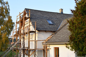 Roof construction with unfinished grey eaves and wooden scaffolding next to renovated white house with asphalt shingles roof in summer day. Home improvement and diy concept.
