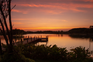 Fototapeta premium Wooden walkway into the lake with natural scenery of sunset and silhouette of forest in background