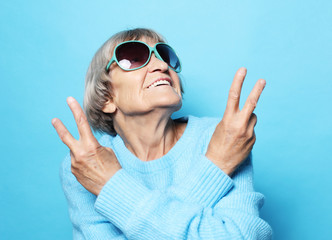 Funny old lady wearing blue sweater, hat and sunglasses showing victory sign