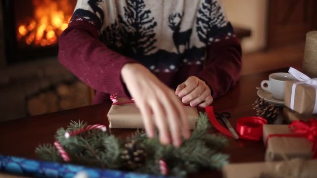 Close view of man's hands tying a bow on a gifts for Christmas holidays near fireplace. Male wrapping present boxes in paper, decorating with fir branches, cones, cane candies on wooden table.
