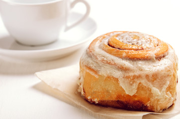 Warm cinnamon bun with icing sugar with cup of coffee on a white table. Sweet pastries, breakfast, snack.