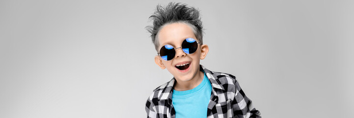 A handsome boy in a plaid shirt, blue shirt and jeans stands on a gray background. The boy is wearing round glasses. The boy is smiling. The boy has an ashy hair color