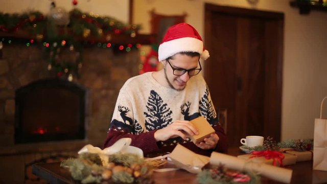 Happy man wearing santa hats opening Christmas gift box near fireplace with flashing garland. Smiling guy recieved a parcel with presents and enjoying unpacking package. Winter holidays concept.