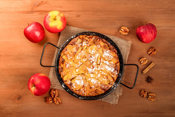 A photo of an apple pie in a pan, shot from above on a dark rustic wooden background with apples, walnuts, cinnamon, anise, and a place for text