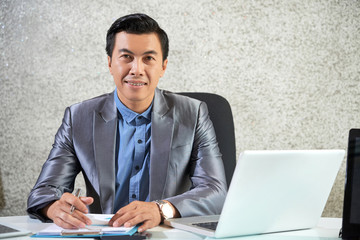 Portrait of smiling male leader in suit sitting at his desk with laptop and signing some documents