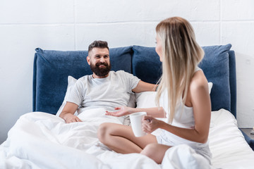 blonde girl with coffee cup talking to smiling bearded man lying in white pillows in bed