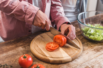 Cropped view of man cutting tomato on wooden board in kitchen