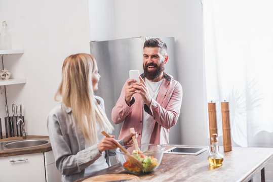 smiling bearded man taking picture of woman at kitchen