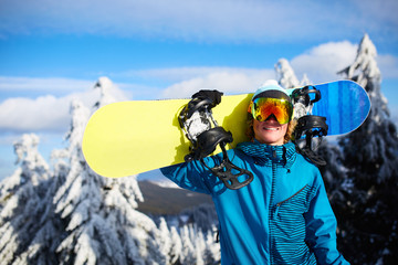 Smiling snowboarder posing carrying snowboard on shoulder at ski resort near forest before...