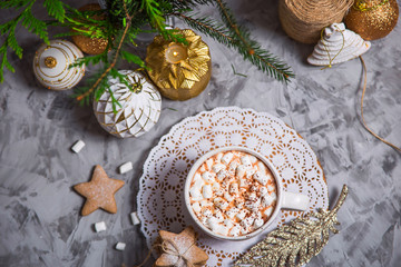 A large cup of cocoa with marshmallow sprinkled with cocoa powder stands on a gray table among Christmas decorations, fir branches, ginger cookies and shiny stars. Top view