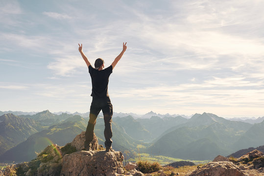 Mann von hinten steht auf dem Gifpel und schaut auf die umliegenden Berge, in blau