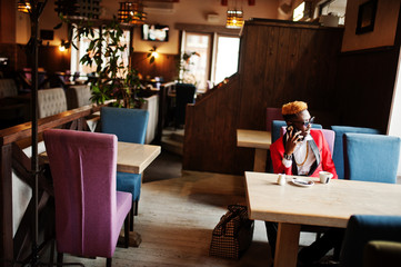 Fashion african american man model at red suit, with highlights hair and sunglasses sitting at cafe with coffee and speaking on mobile phone.