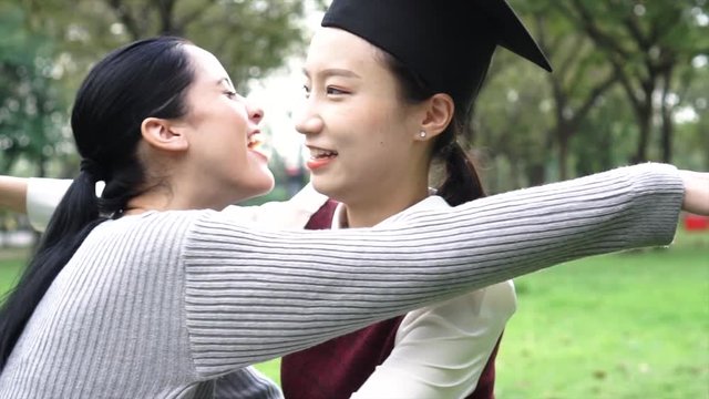 Young Asian female graduate hugging her friend at graduation ceremony in slow motion. International diversity background