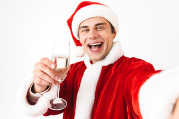 Portrait of handsome man 30s in santa claus costume and red hat taking selfie photo while holding glass with champagne, isolated over white background in studio