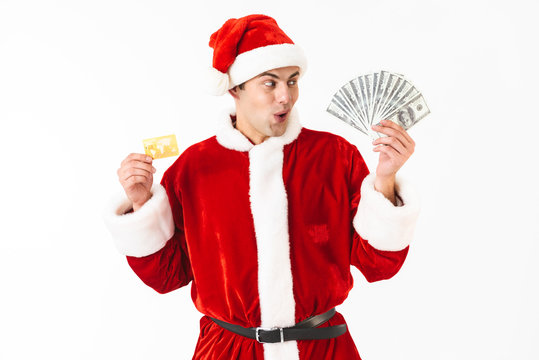 Image of surprised man 30s in santa claus costume holding dollar bills and credit card, isolated on white background in studio