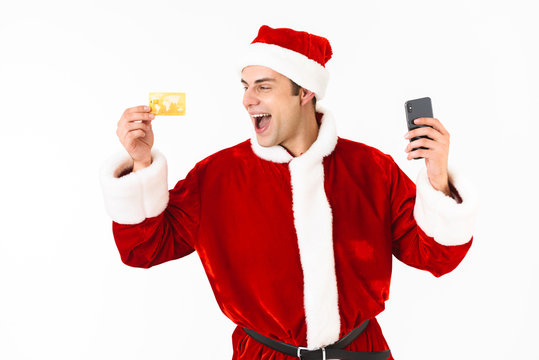 Image of optimistic man 30s in santa claus costume holding cell phone and credit card, isolated on white background in studio