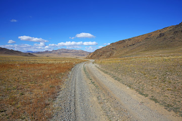 Fototapeta na wymiar The rural road in the desert behind the mountain range with sky blue and cluods. Central Asia between the Russian Altai and Western Mongolia 