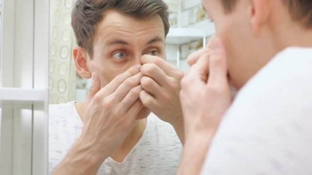 Good looking young man standing close to a mirror in the bathroom and squeezing a pimple on his face.