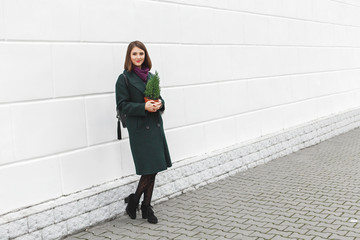 Beautiful young stylish brunette woman wearing green coat walking through the city streets with small Christmas tree in her hands. New year concept. Christmas tree in a pot.