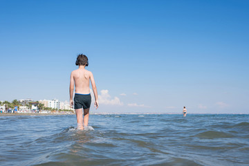 Happy cheerful teen boy on the beach with the headphones running on the wave of the sea on the resort town background. Concept