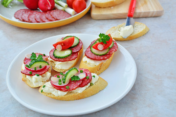 Tasty appetizers with cream cheese, salami, red radish, scallions, cucumbers and tomatoes on white plate over marbled counter top.