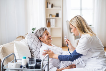 A health visitor examining a sick senior woman lying in bed at home with stethoscope.