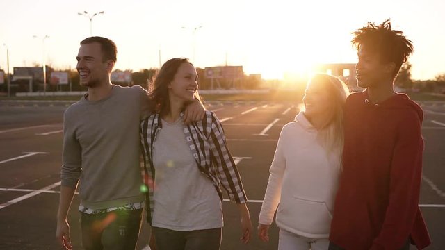 Group of four young multirace friends laughing while walking outdoors. Young men and women enjoying a summer day at parking zone near the mall. Evening dusk