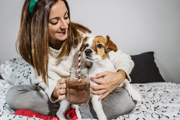 .Young and cheerful woman playing with her nice dog on Christmas morning, drinking a hot chocolate. Both with christmas costume. Lifestyle.
