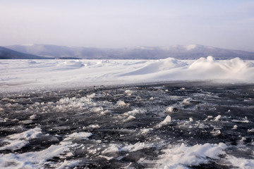 Natural winter lake ice breaking with clear sky background, selective focus. Frozen mountain lake with blue ice and cracks on the surface. Winter landscape with snowy hills under a blue sky.
