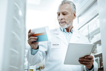 Pleased gray-haired pharmacist reading ingredients on box