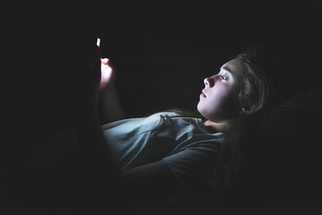 Depressed girl lying down on a couch in the dark while using her smartphone. The light from the...
