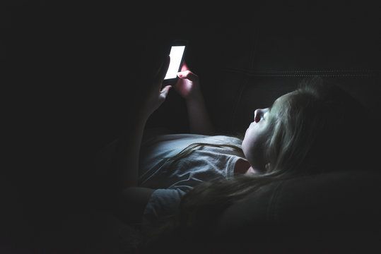 Depressed girl lying down on a couch in the dark while using her smartphone. The light from the screen is illuminating her face.