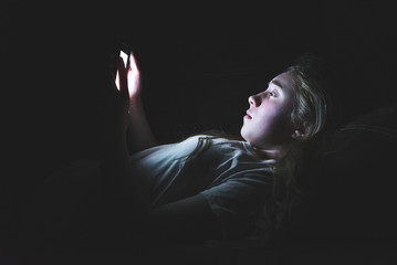 Depressed girl lying down on a couch in the dark while using her smartphone. The light from the...