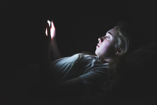 Depressed girl lying down on a couch in the dark while using her smartphone. The light from the screen is illuminating her face.