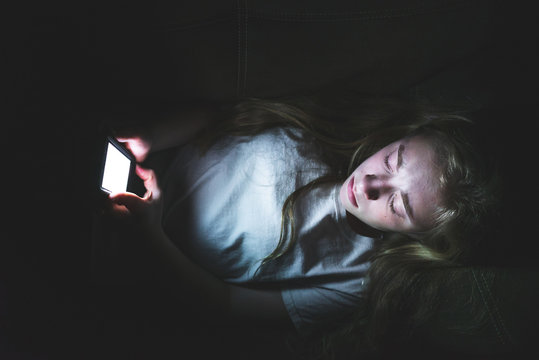 Depressed Girl Lying Down On A Couch In The Dark While Using Her Smartphone. The Light From The Screen Is Illuminating Her Face.