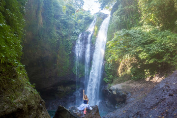 Young woman with long hair sitting on the rock in front of Aling-Aling waterfall among green tropical trees and plants on the north of Bali island, Indonesia