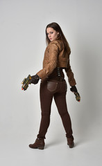 full length portrait of brunette  girl wearing brown leather steampunk outfit. standing pose, holding a gun, on grey studio background.
