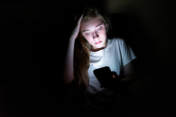 Upset girl sitting in the dark while using her smartphone. The light from the screen is illuminating her face.