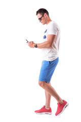 Fototapeta na wymiar Side view of young man in summer clothes walking and using mobile phone. Full body isolated on white background. 