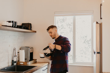 Young Attractive Minimalist Hipster Man in Modern Designed Kitchen Brews, Pours, and Drinks Morning Coffee out of a Cup