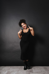 Full length photo of pretty african american woman wearing black dress gesturing, isolated over dark background