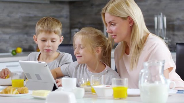 Happy little boy and girl laughing and playing on digital tablet while having breakfast together with mom