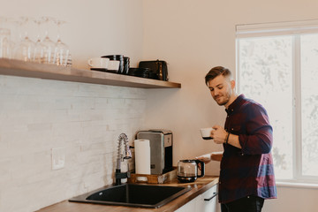 Young Attractive Minimalist Hipster Man in Modern Designed Kitchen Brews, Pours, and Drinks Morning Coffee out of a Cup