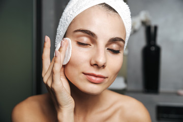 Photo of pretty woman with towel on head cleaning her face and removing makeup with cotton pad, while standing in bathroom