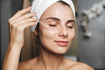 Photo of lovely woman with towel on head applying cosmetic oil on her face, while standing in bathroom
