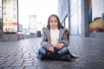 Child girl is sitting and smiling in the middle of the sidewalk in evening.